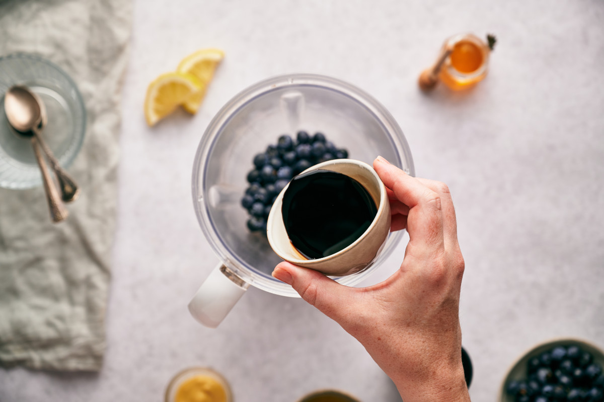 Woman's hand pouring balsamic vinegar into a blender full of blueberries.