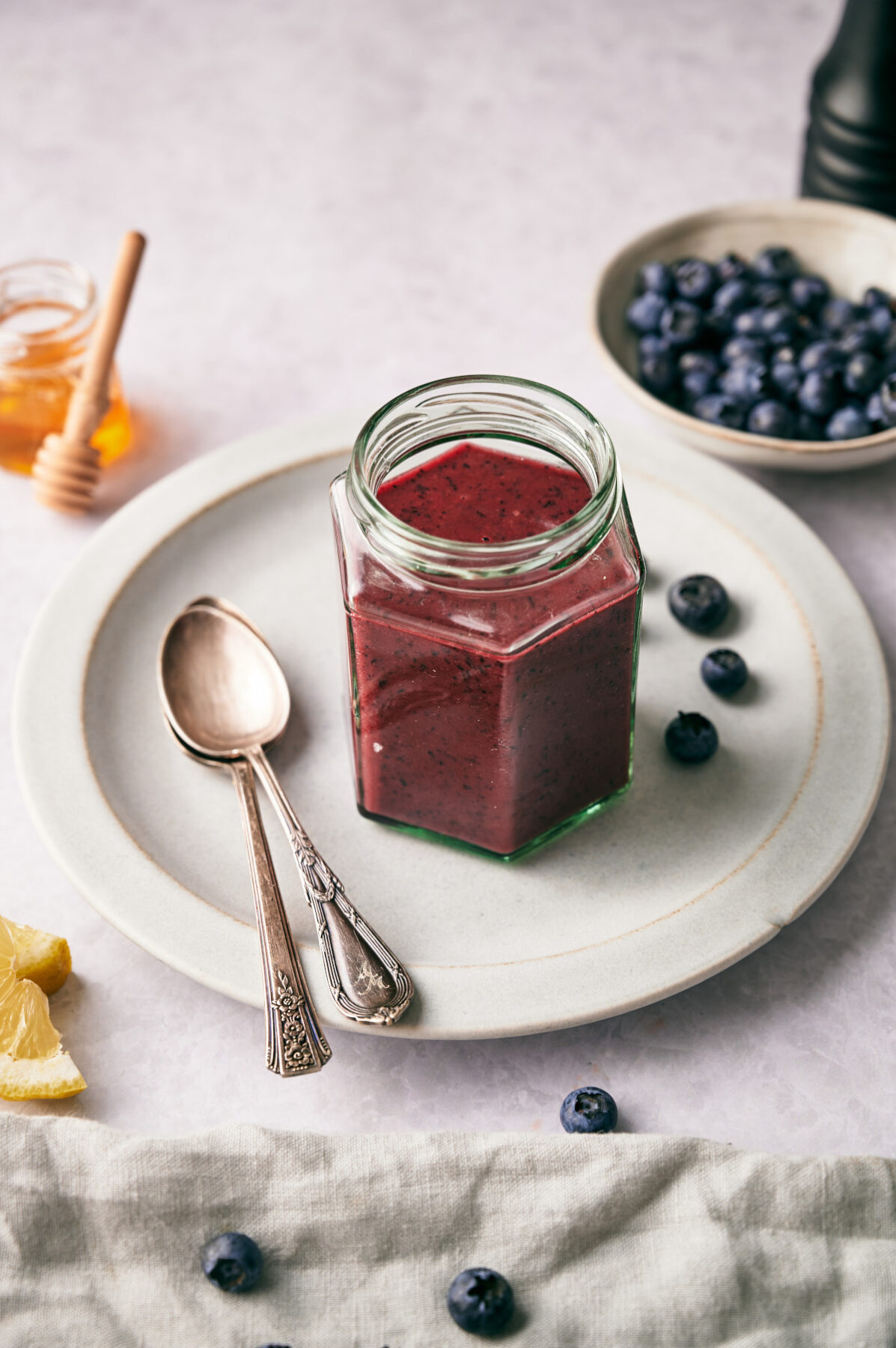Blueberry Vinaigrette dressing in a glass jar on a white plate with two spoons, blueberries and honey in the background.