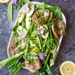top down cropped photo of a serving dish containing grilled spring vegetables including french green beans, spring onions, sugar snap peas, asparagus, leeks, and artichokes, glisten with a homemade lemon parsley butter